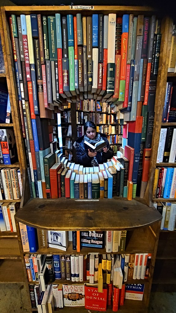 Los Angeles literary tour. South Asian woman wears scarf and puffer jacket while reading a book through large peephole surrounded by books at the Last Bookstore